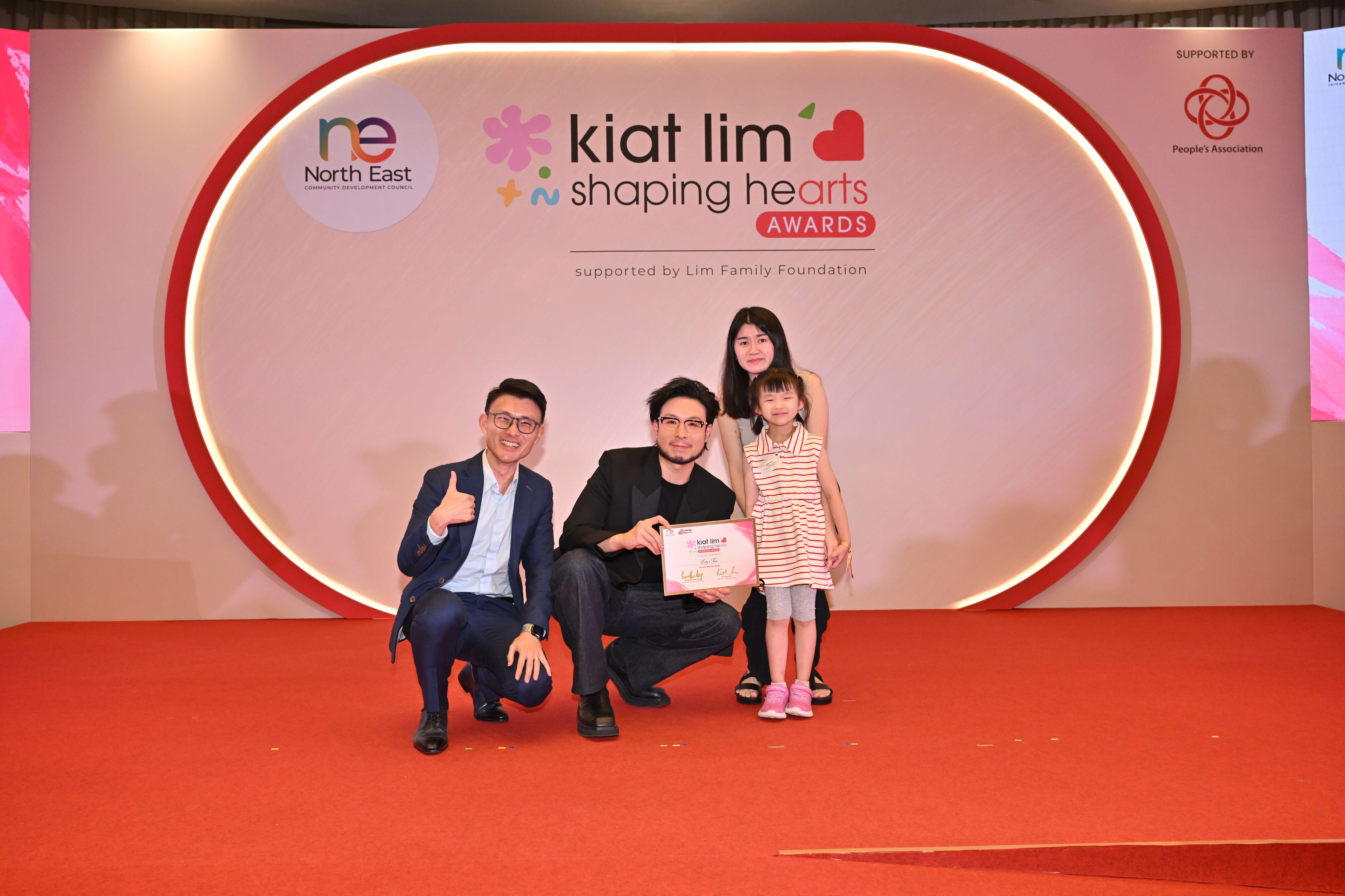 Award recipient standing on stage with the presenter, holding their certificate and posing for a group photo during the award ceremony, with a backdrop reading ‘Kiat Lim-Shaping Hearts Awards'.